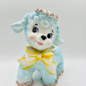 Puede incluir: Un macetero de cerámica azul con forma de cordero con un lazo amarillo. El cordero tiene una cara blanca con ojos negros y una nariz rosa. El macetero tiene una corona de flores y un borde rosa y blanco alrededor de la base.