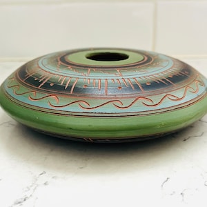 May include: A green ceramic bowl with a circular design in black, blue, and brown. The bowl has a small hole in the center.
