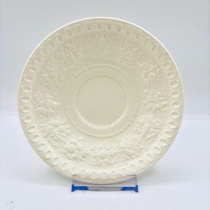 May include: Cream-coloured vintage saucer with embossed floral and fruit designs. The rim features a repeating pattern of small circles. The centre has a recessed area. A decorative piece for serving or display.