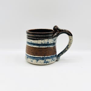 May include: Handmade ceramic mug with a unique handle design. The mug features horizontal bands of brown, blue, and white, with a speckled texture. The interior and rim are dark brown. A rustic, handcrafted piece for enjoying beverages.