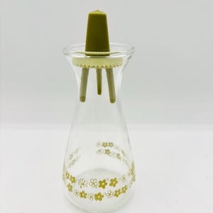 Puede incluir: Aceitera de vidrio transparente con tapón de plástico verde y estampado floral. El tapón tiene una parte superior cónica y tres patas. La base de la aceitera presenta un patrón repetido de pequeñas flores y hojas verdes.