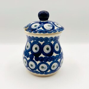 May include: A ceramic jar with a lid, featuring a cream-coloured base decorated with a repeating pattern of blue and white circular eye motifs. The jar has a rounded body and a small, dark blue knob handle on the centre of the lid.