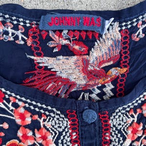 May include: Close-up of a navy blue top with intricate embroidery. The design features a large bird with red, white, and orange details, along with floral patterns. The tag reads "JOHNNY WAS".