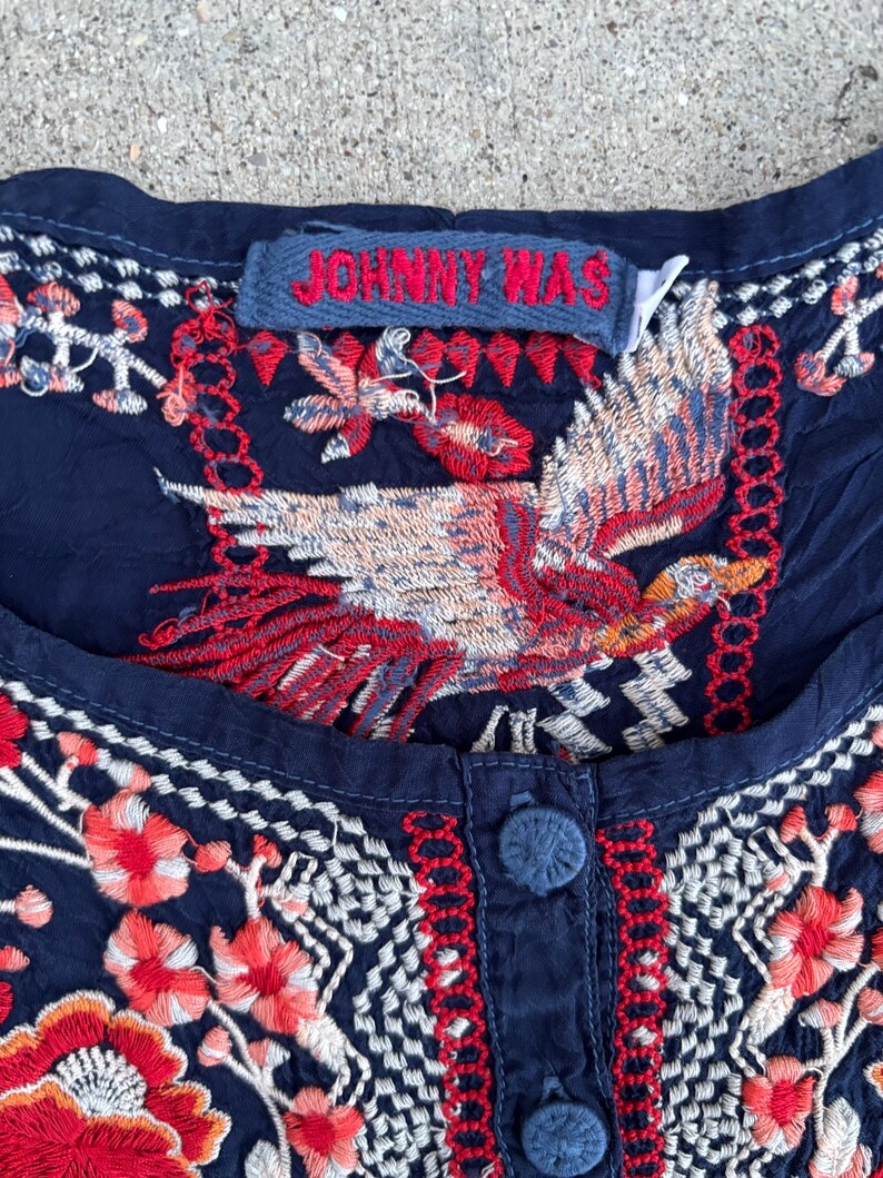 May include: Close-up of a navy blue top with intricate embroidery. The design features a large bird with red, white, and orange details, along with floral patterns. The tag reads "JOHNNY WAS".