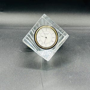 May include: A clear crystal clock with a square shape. The clock face is white with black Roman numerals and gold trim. The words "WATERFORD CRYSTAL" are printed on the clock face.