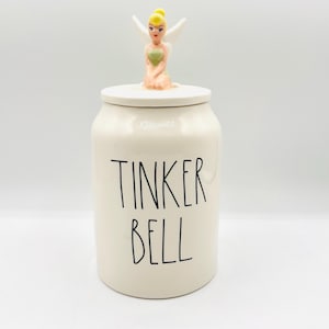 Może przedstawiać: Kremowy ceramiczny słoik z pokrywką, z czarnym napisem "TINKER BELL". Figurka Dzwoneczka z żółtymi włosami, białymi skrzydłami oraz zieloną i różową sukienką siedzi na pokrywce. Słoik ma cylindryczny kształt.