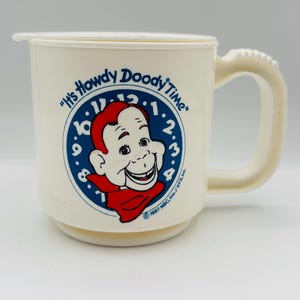 Puede incluir: Taza de cerámica blanca con una ilustración azul y roja de Howdy Doody, un personaje de televisión infantil. La taza tiene un asa y dice "It's Howdy Doody Time" en la parte delantera. © 1987 NBC Inc./KFS Inc.