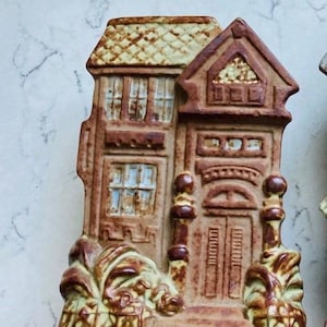 May include: A brown ceramic house-shaped ornament with a detailed facade, including windows, a door, and a textured roof. The ornament has a vintage aesthetic, with a decorative base and a light-colored roof.
