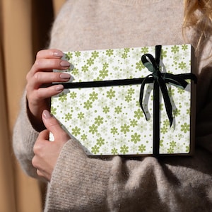 Könnte beinhalten: Eine Geschenkbox, verpackt in weißem Papier mit einem Muster aus grünen Blumen und einem schwarzen Samtband. Die Box wird von einer Person gehalten, die einen beigen Pullover trägt.