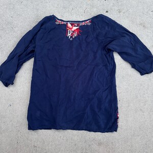 May include: A navy blue long-sleeve top with embroidered details. The neckline features red, white, and black floral embroidery. The top has a relaxed fit and is made of a soft, flowing fabric. The sleeves are three-quarter length.