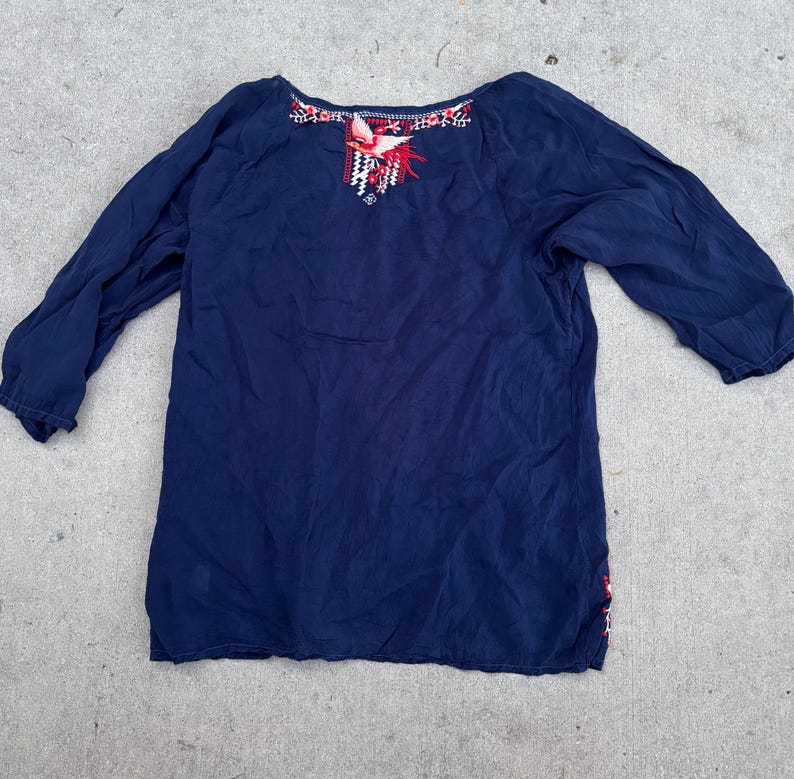 May include: A navy blue long-sleeve top with embroidered details. The neckline features red, white, and black floral embroidery. The top has a relaxed fit and is made of a soft, flowing fabric. The sleeves are three-quarter length.