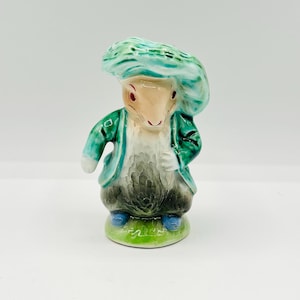 May include: A ceramic figurine of a rabbit-like character wearing a teal hat and jacket. The figure has a white chest, gray and black pants, and blue shoes. It stands on a green base.