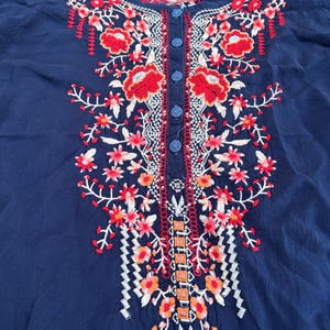 May include: Navy blue blouse with intricate floral embroidery in red, orange, and white. The embroidery is concentrated around the neckline and down the front, with small blue buttons. The blouse has a round neckline and short sleeves.