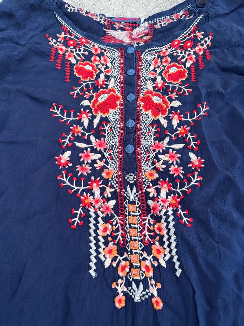 May include: Navy blue blouse with intricate floral embroidery in red, orange, and white. The embroidery is concentrated around the neckline and down the front, with small blue buttons. The blouse has a round neckline and short sleeves.