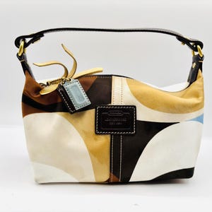 May include: A Coach handbag with a brown leather handle and a colorful geometric pattern in shades of brown, tan, white, and blue. The bag has a small leather tag and a rectangular patch with the brand name.