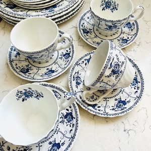 Puede incluir: Juego de té de porcelana blanca con un diseño floral azul. El conjunto incluye varias tazas, platillos y platos, dispuestos sobre una superficie blanca. El diseño azul presenta motivos florales y bordes decorativos.