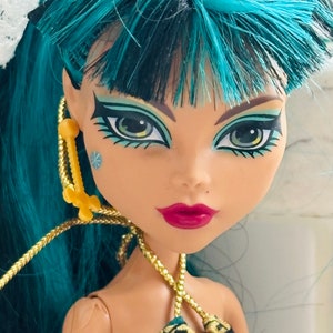 Peut inclure: Une poup&eacute;e avec des cheveux bleu sarcelle et noirs, portant un haut &agrave; motifs noirs et dor&eacute;s. La poup&eacute;e a la peau brune, des yeux bleus avec un eye-liner noir et des l&egrave;vres roses.
