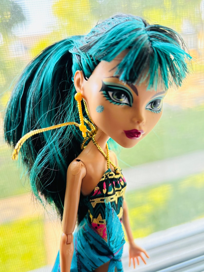Peut inclure: Une poup&eacute;e aux cheveux bleu turquoise portant des v&ecirc;tements &agrave; motifs noirs et dor&eacute;s. La poup&eacute;e a les yeux bleus et un maquillage fonc&eacute;. La poup&eacute;e porte des boucles d'oreilles dor&eacute;es.