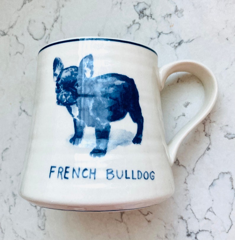 May include: White ceramic mug with a blue illustration of a French Bulldog. The mug has a curved handle and the words "FRENCH BULLDOG" are printed in blue below the dog image. The rim of the mug is trimmed with a thin blue line.
