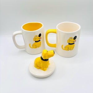 Puede incluir: Dos tazas de cerámica blanca con interior amarillo y una ilustración de perro de dibujos animados. Una taza tiene un asa amarilla, la otra un asa blanca. Una figura de perro amarillo con collar negro sobre una base redonda blanca.