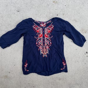 May include: A navy blue blouse with three-quarter sleeves. The blouse features intricate floral embroidery in red, pink, and white down the front and on the sleeves. The embroidery is detailed and adds a decorative element to the top.