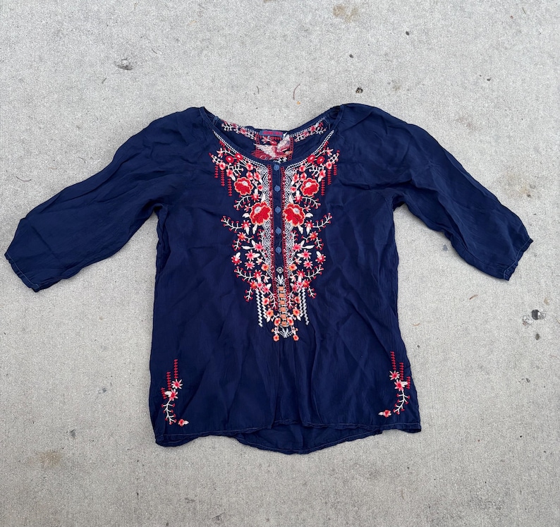 May include: A navy blue blouse with three-quarter sleeves. The blouse features intricate floral embroidery in red, pink, and white down the front and on the sleeves. The embroidery is detailed and adds a decorative element to the top.