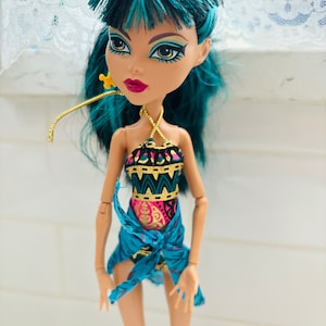 Peut inclure: Une poup&eacute;e aux cheveux turquoise portant un maillot de bain &agrave; motifs bleus et dor&eacute;s avec un sarong bleu. La poup&eacute;e a les yeux verts et le rouge &agrave; l&egrave;vres rouge.
