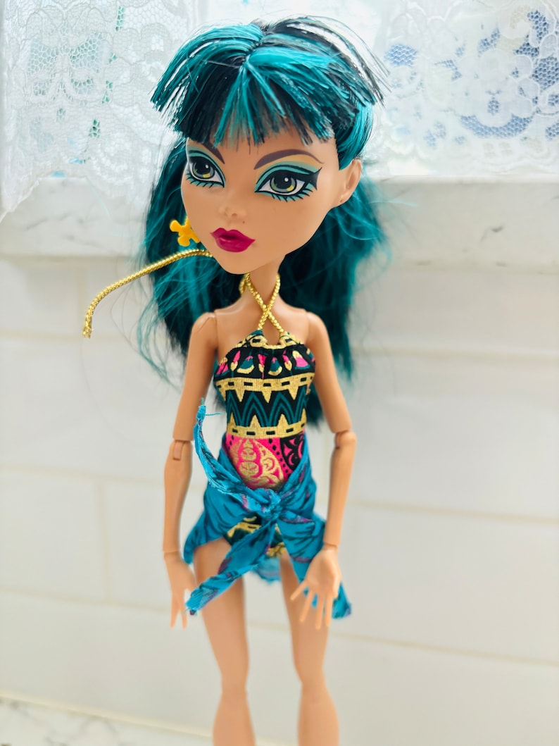 Peut inclure: Une poup&eacute;e aux cheveux turquoise portant un maillot de bain &agrave; motifs bleus et dor&eacute;s avec un sarong bleu. La poup&eacute;e a les yeux verts et le rouge &agrave; l&egrave;vres rouge.