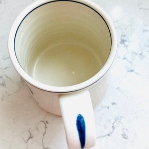 May include: A white ceramic mug with a blue accent on the handle and a thin blue line around the rim. The mug has a simple, minimalist design, perfect for coffee or tea.
