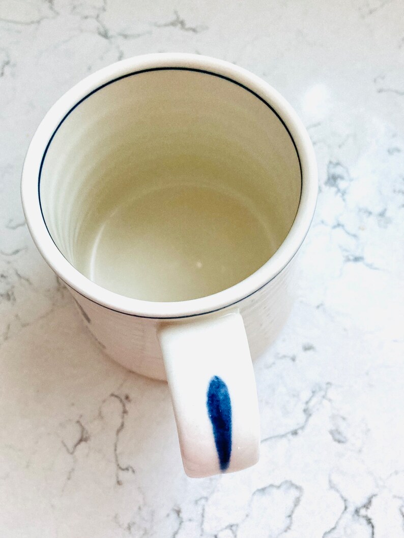 May include: A white ceramic mug with a blue accent on the handle and a thin blue line around the rim. The mug has a simple, minimalist design, perfect for coffee or tea.
