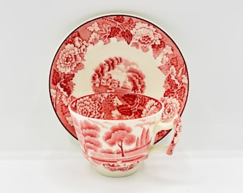 Può includere: Un set di tazza e piattino rosso e bianco. La tazza e il piattino presentano un design floreale e paesaggistico. Il piattino ha un bordo floreale e una scena paesaggistica centrale. La tazza ha una scena paesaggistica abbinata e un manico decorativo.