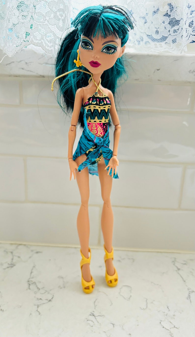 Peut inclure: Une poup&eacute;e aux cheveux turquoise portant un maillot de bain bleu et or avec un par&eacute;o assorti. La poup&eacute;e a un maquillage fonc&eacute; et porte des talons hauts jaunes.