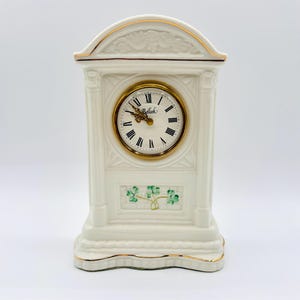 Puede incluir: Un reloj de porcelana blanca con detalles dorados y un diseño de trébol verde. La esfera del reloj tiene números romanos y la palabra "Belleck" impresa en ella.