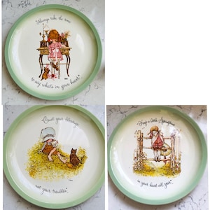 Puede incluir: Conjunto de tres platos de cerámica con borde verde y fondo blanco. Cada plato presenta una ilustración diferente de una niña con un gato y una cita. El primer plato dice "Siempre tómate el tiempo para decir lo que está en tu corazón". El segundo plato dice "Cuenta tus bendiciones, no tus problemas". El tercer plato dice "Mantén un poco de primavera en tu corazón todo el año".