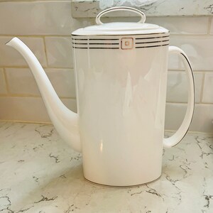 Puede incluir: Cafetera de cerámica blanca con ribete dorado y diseño cuadrado. La cafetera tiene una tapa con asa y un pico.