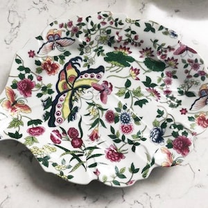 May include: A white ceramic serving dish shaped like a leaf with a floral and butterfly design. The dish features pink, yellow, blue, and green flowers and butterflies.