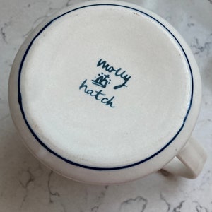 May include: A white ceramic mug with a blue rim and handle. The bottom of the mug features the handwritten text "molly hatch" in dark blue. The mug is on a white and gray surface.