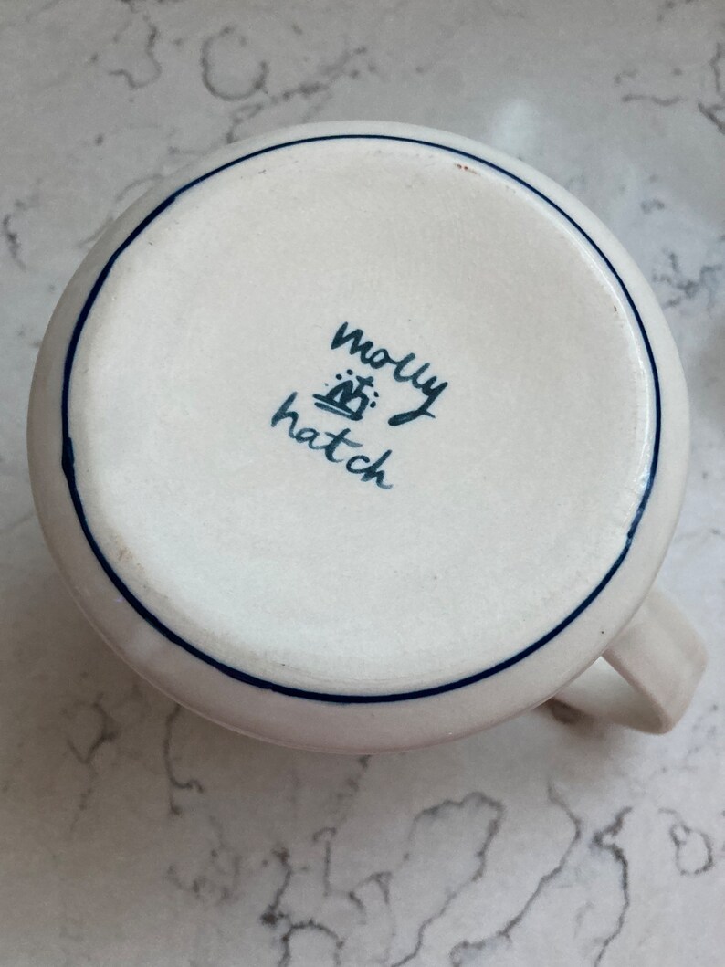 May include: A white ceramic mug with a blue rim and handle. The bottom of the mug features the handwritten text "molly hatch" in dark blue. The mug is on a white and gray surface.