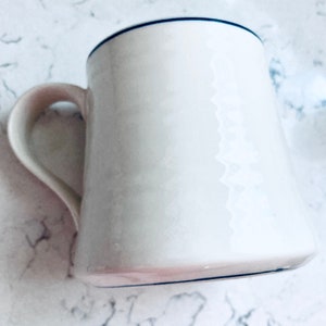 May include: White ceramic mug with a curved handle and a dark blue rim. The mug has a slightly flared shape and a smooth, glossy finish. The mug is empty and sits on a white marble surface.