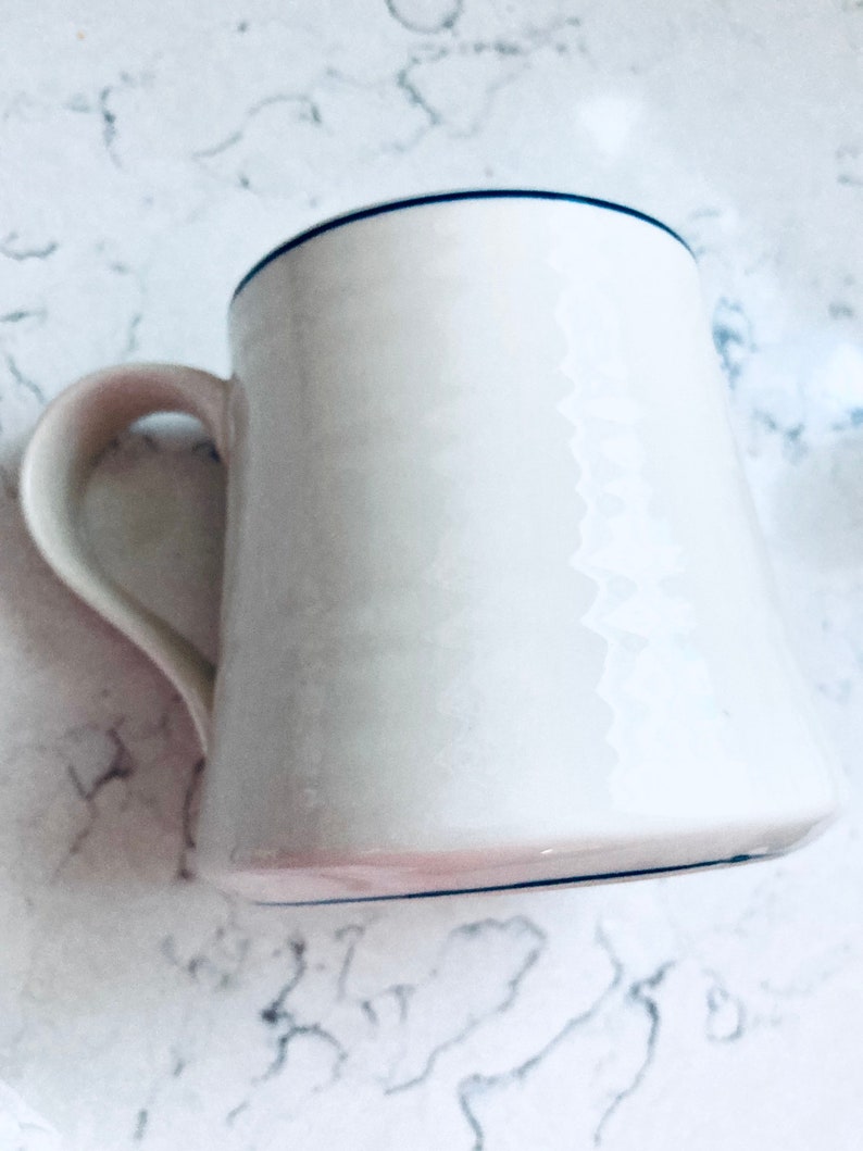 May include: White ceramic mug with a curved handle and a dark blue rim. The mug has a slightly flared shape and a smooth, glossy finish. The mug is empty and sits on a white marble surface.