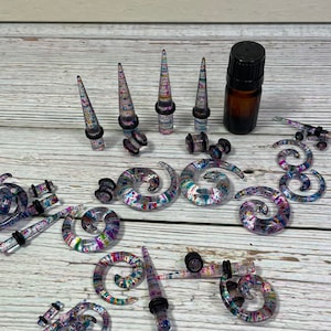 May include: A set of colorful spiral and spike ear gauges made of clear acrylic with black o-rings. The gauges are scattered on a white wooden surface. A small bottle of oil is also included.