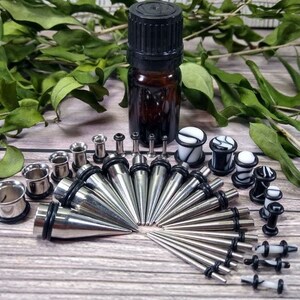 May include: A set of body jewelry featuring a variety of ear gauges, plugs, and tapers. The set includes a mix of silver, black, and white colors. The jewelry is arranged on a wooden surface.