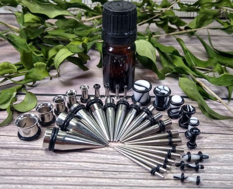 May include: A set of body jewelry featuring a variety of ear gauges, plugs, and tapers. The set includes a mix of silver, black, and white colors. The jewelry is arranged on a wooden surface.