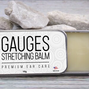 May include: A silver tin with a white label that reads "GAUGES STRETCHING BALM PREMIUM EAR CARE 10g MADE IN USA". The tin is open and a white balm is visible inside.