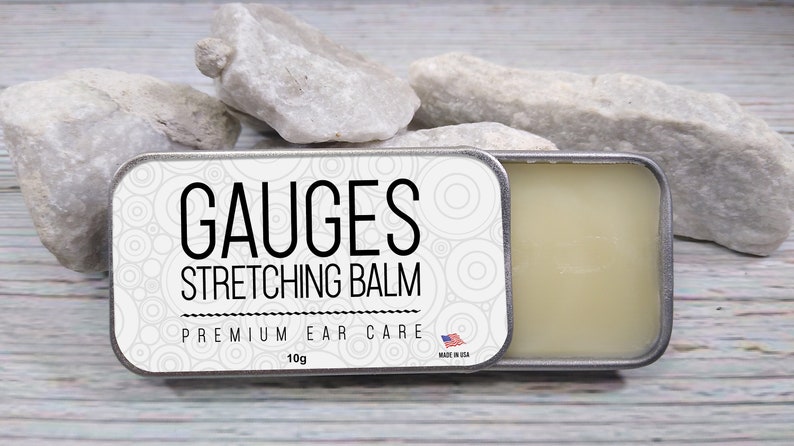 May include: A silver tin with a white label that reads "GAUGES STRETCHING BALM PREMIUM EAR CARE 10g MADE IN USA". The tin is open and a white balm is visible inside.