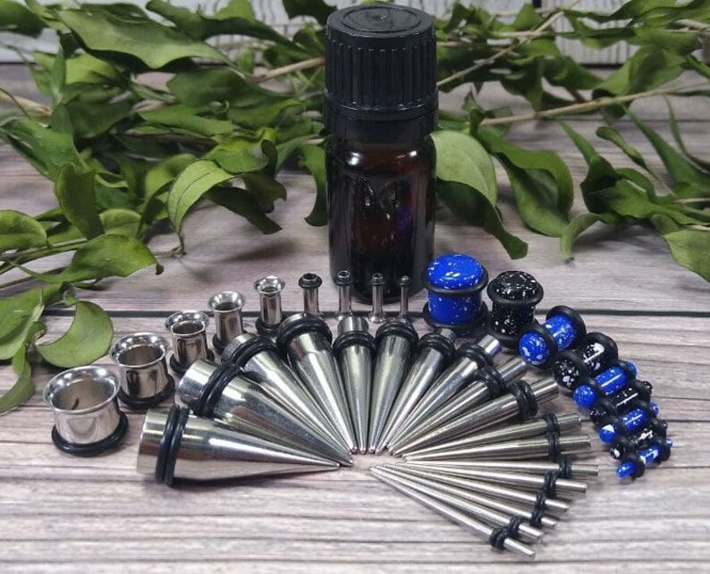 May include: A set of 16 stainless steel ear gauges, including a variety of sizes and styles. The set includes a small bottle of oil, likely for cleaning and lubrication. The gauges are a mix of plain and patterned, with some featuring a blue and white speckled design.