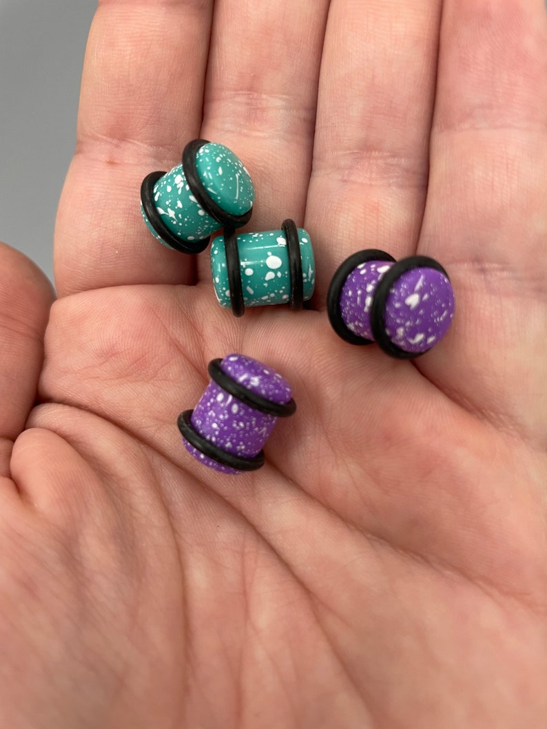 May include: Four pairs of ear gauges with black o-rings. Two pairs are teal with white speckles and two pairs are purple with white speckles.
