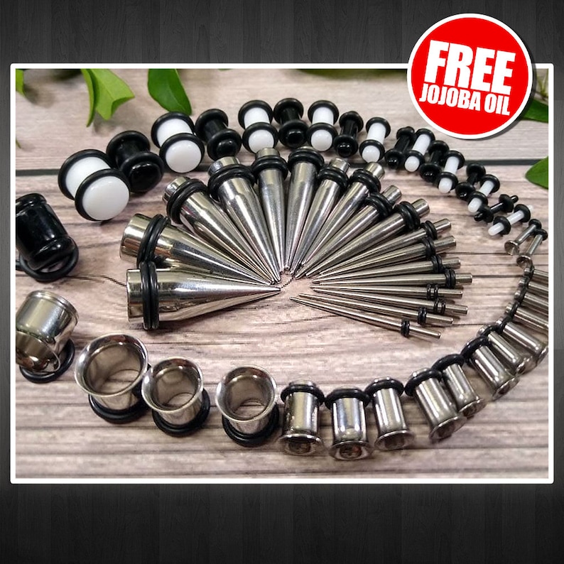 May include: A set of silver and black ear gauges and tapers. The set includes a variety of sizes and styles, including plugs, tunnels, and tapers. The tapers are silver and have a pointed tip. The plugs and tunnels are silver with a black rubber o-ring. The set is displayed on a wooden surface. FREE JOJOBA OIL is written on a red circle.