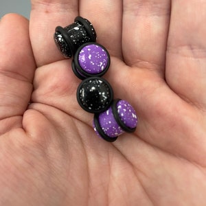 May include: Four pairs of ear gauges, two black with white speckles and two purple with white speckles. The gauges are made of acrylic and have a black rubber o-ring.