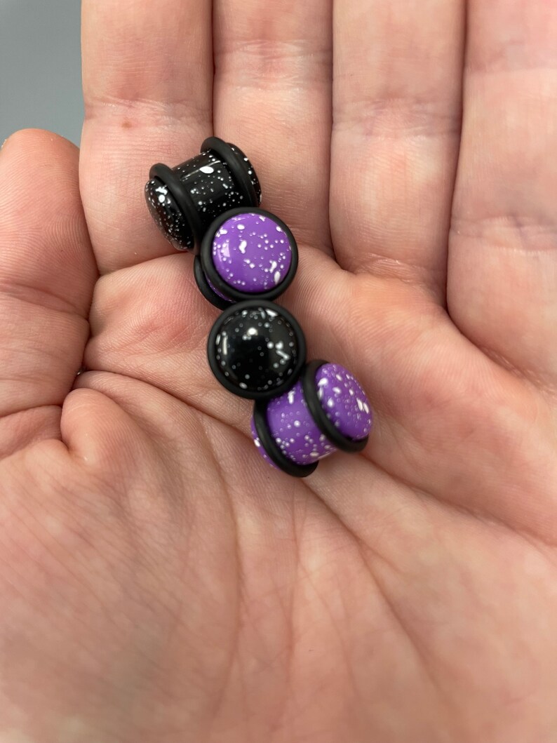 May include: Four pairs of ear gauges, two black with white speckles and two purple with white speckles. The gauges are made of acrylic and have a black rubber o-ring.